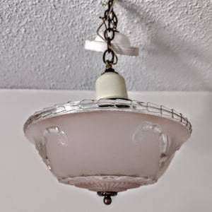 May include: A vintage ceiling light fixture with a frosted pink glass shade. The shade has a decorative etched design and is trimmed with a silver-colored metal. The fixture hangs from a chain and a brass-colored canopy.