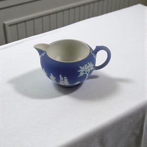 May include: A small, blue creamer with a white interior and handle. The exterior features white relief designs of figures and trees. The creamer is likely made of ceramic or porcelain and is set on a white surface.