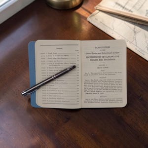May include: An open vintage booklet titled Constitution of the Grand Lodge and Subordinate Lodges Brotherhood of Locomotive Firemen and Enginemen with a contents page on the left and text on the right, featuring a silver pen resting across the pages.