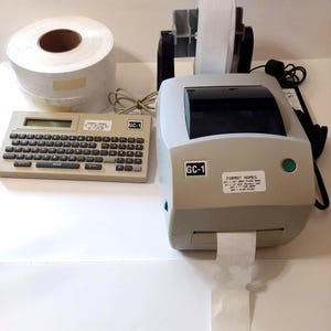 以下が含まれることがあります： キーボード、ラベルロール、プリンターを備えたラベルメーカー。 ラベルメーカーはベージュ色で、黒いキーボードと「GC-1」の文字があります。 プリンターはグレーとベージュで、「GC-1」と「FORMAT NAMES」の文字があります。