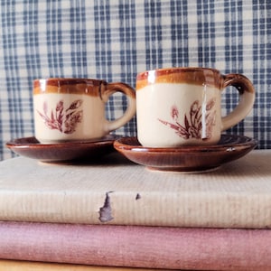 May include: Two vintage espresso cups and saucers. The cups are off-white with a brown rim and handle, and a maroon floral design. The saucers are brown. The cups and saucers are sitting on top of a stack of books.