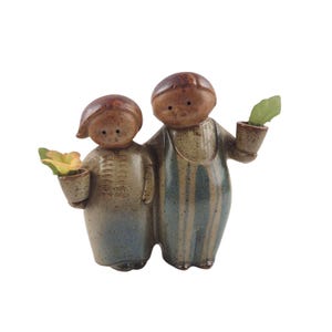 Pode incluir: Uma estatueta de cerâmica retratando duas figuras, uma com macacão listrado azul, a outra com uma roupa azul claro e bege. Cada figura segura um pequeno vaso com uma planta. A estatueta tem uma aparência rústica e artesanal.