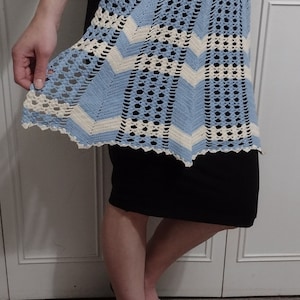May include: A blue and white crocheted apron with a scalloped edge. The apron is worn over a black skirt.