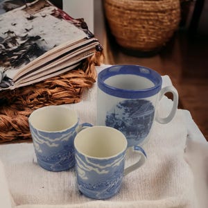 May include: Three blue and white ceramic mugs with a blue rim. The mugs have a blue and white design with a scene of a building and trees. The mugs are sitting on a white surface.