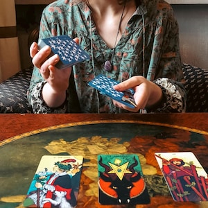 May include: A person holding a deck of blue star-patterned tarot cards, with three tarot cards displayed on a table. The cards feature colorful illustrations, including a figure in red and a black horned creature.