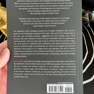 May include: A black and white book cover with the title "Wanderer's Tarot Guidebook" and the author's name "Casey Zabala". The cover features a quote from the book, "Wanderer's Tarot draws you in like nobody's business. It's as though this deck does a very quiet, very subtle spell on you. It draws a veil across your eyes, then unravels it, thread by thread." -Beth Maiden, Little Red Tarot