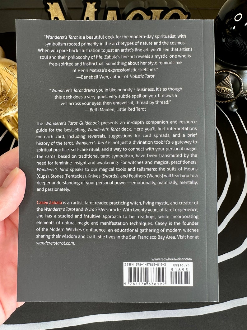 May include: A black and white book cover with the title "Wanderer's Tarot Guidebook" and the author's name "Casey Zabala". The cover features a quote from the book, "Wanderer's Tarot draws you in like nobody's business. It's as though this deck does a very quiet, very subtle spell on you. It draws a veil across your eyes, then unravels it, thread by thread." -Beth Maiden, Little Red Tarot