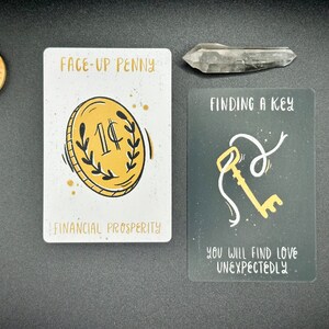 May include: Two fortune telling cards. The first card is white with a gold penny and the text "FACE-UP PENNY" and "FINANCIAL PROSPERITY". The second card is black with a gold key and the text "FINDING A KEY" and "YOU WILL FIND LOVE UNEXPECTEDLY".
