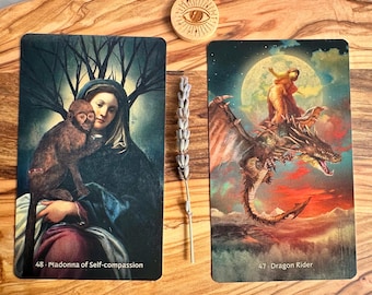 May include: Two tarot cards, "48: Madonna of Self-compassion" and "47: Dragon Rider", are displayed on a wooden surface. A sprig of lavender and a wooden disc with an eye symbol are also present. The cards feature detailed illustrations with vibrant colors.