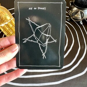 May include: A black tarot card with white text and a white illustration of a crystal. The card reads "Ace of Stones".