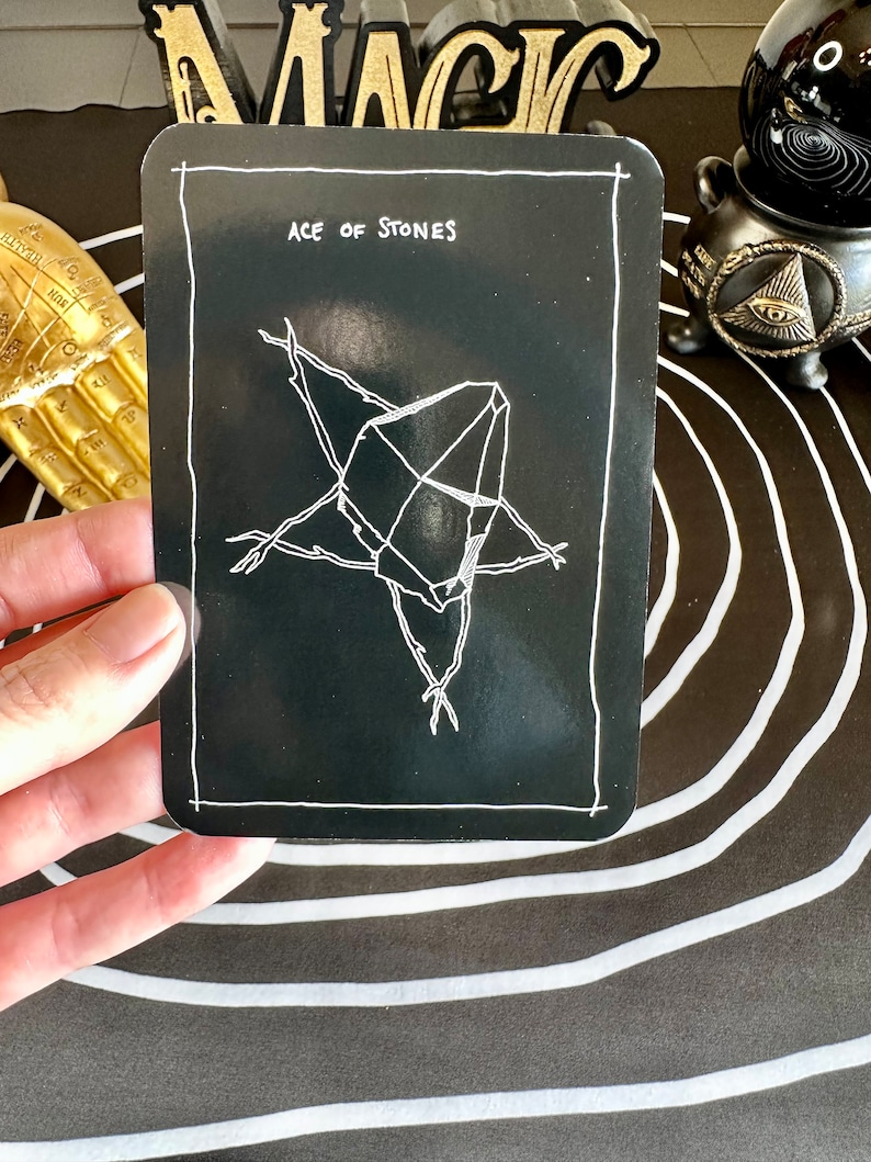 May include: A black tarot card with white text and a white illustration of a crystal. The card reads "Ace of Stones".