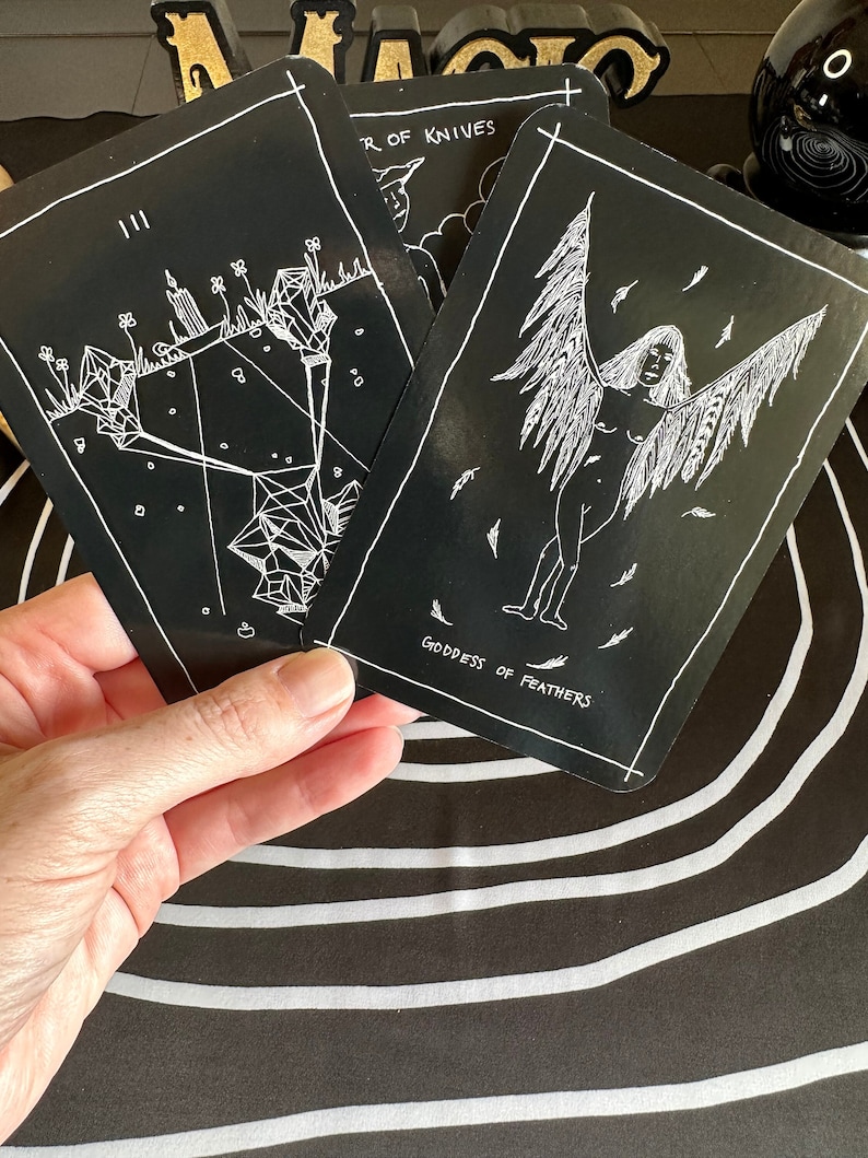 May include: Two black tarot cards with white illustrations. The card on the left is the Three of Swords, depicting a crystal structure with three candles. The card on the right is the Goddess of Feathers, showing a figure with wings and feathers.