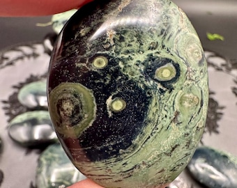 May include: An oval-shaped, polished stone with a dark green and black swirling pattern. The stone has lighter green circular patterns and a glossy finish. The stone is held by a person with blue nail polish.
