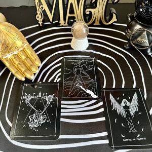 May include: Three black tarot cards with white illustrations on a black and white circular background. The cards are "Philosopher of Knives", "III", and "Goddess of Feathers".