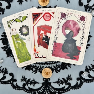 May include: Three tarot cards with illustrations and text. The cards are labeled "17. MIRROR", "18. FEVER", and "19. MOURNING". The "MIRROR" card features a woman in green, the "FEVER" card shows a red bed, and the "MOURNING" card depicts a woman in black. Two wooden eye-shaped tokens are also visible.
