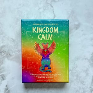 May include: A colourful box titled "KINGDOM CALM" featuring a winged cat illustration. The box has a rainbow gradient and text stating "56 Practices for Children (and those who love them) to support nervous system regulation & emotional well-being."