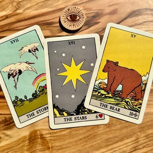May include: Three tarot cards with illustrations of storks, a star, and a bear, and a wooden disc with an eye symbol. The cards have white borders and feature the titles "The Stork", "The Stars", and "The Bear". The background is a wooden surface.