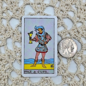 May include: A tarot card featuring a figure holding a golden cup, with a blue and red outfit, standing on yellow ground with blue waves. The card is labeled "PAGE of CUPS."