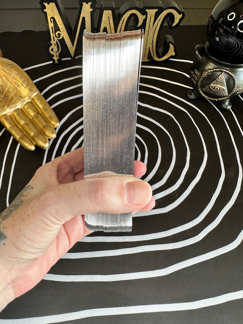 May include: A stack of silver playing cards, held in a hand, against a black and white background with a circle pattern.