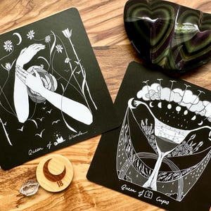 Może przedstawiać: Dwie czarne karty tarota z białymi ilustracjami, jedna z dłońmi i napisem "Queen of Wands", druga z wzorem kubka z napisem "Queen of Cups". Obecny jest r&oacute;wnież kamień w kształcie serca w kolorze zielono-fioletowym oraz drewniany żeton w kształcie księżyca.