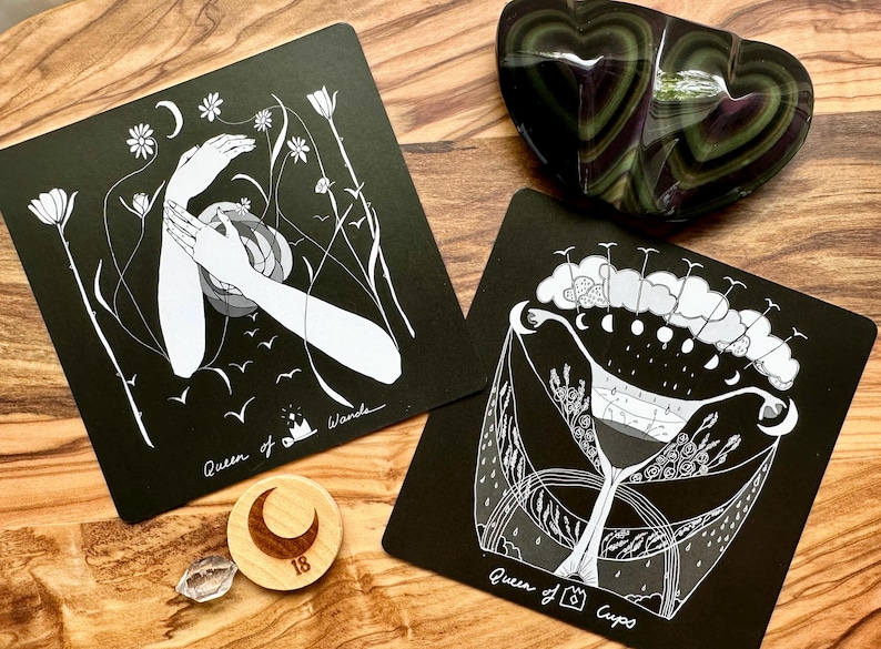 Może przedstawiać: Dwie czarne karty tarota z białymi ilustracjami, jedna z dłońmi i napisem "Queen of Wands", druga z wzorem kubka z napisem "Queen of Cups". Obecny jest r&oacute;wnież kamień w kształcie serca w kolorze zielono-fioletowym oraz drewniany żeton w kształcie księżyca.