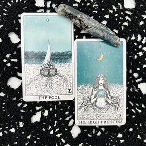 May include: Two tarot cards, "The Fool" and "The High Priestess", with illustrations of a sailboat and a woman in water, respectively. A gray crystal rests diagonally across the cards. The cards are on a black, lacy surface.