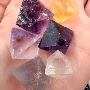 Puede incluir: Una colección de seis cristales pequeños, facetados y con forma de octaedro en varios colores, incluyendo morado oscuro, lavanda claro, transparente y amarillo, cada uno con fracturas internas naturales y superficies translúcidas.