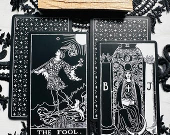 May include: Two black and white tarot cards titled The Fool and The High Priestess, featuring detailed illustrations and white text. A rectangular piece of light brown wood rests horizontally above the cards, which are arranged on a white lace fabric.