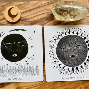 Może przedstawiać: Dwie karty tarota z czarno-białymi ilustracjami Księżyca i Słońca. Karta Księżyca przedstawia twarz, postacie i drzewa. Karta Słońca ma twarz otoczoną kwiatowym wzorem. Obecny jest r&oacute;wnież drewniany żeton z p&oacute;łksiężycem i numerem 18.