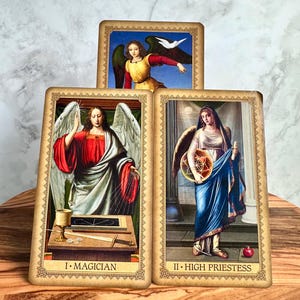 May include: Three tarot cards featuring angel illustrations. The cards have a vintage aesthetic with detailed artwork and gold borders. The cards are labeled "I. MAGICIANS" and "II. HIGH PRIESTESS". The top card shows an angel with a white dove.