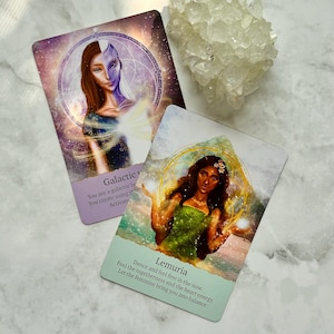 May include: Two oracle cards featuring illustrations of women and text. One card reads "Galactic" and the other "Lemuria". A cluster of clear crystals is in the background. The cards have a celestial theme with purple, blue, and green colours.