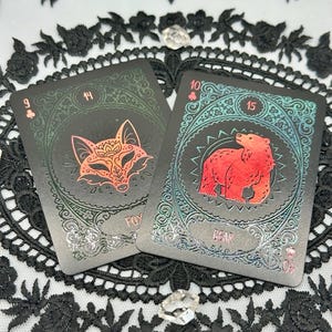 May include: Two tarot cards with animal illustrations. One card features a fox mask, the other a red bear. Both cards have decorative borders and numbers. The cards are set against a black lace background.