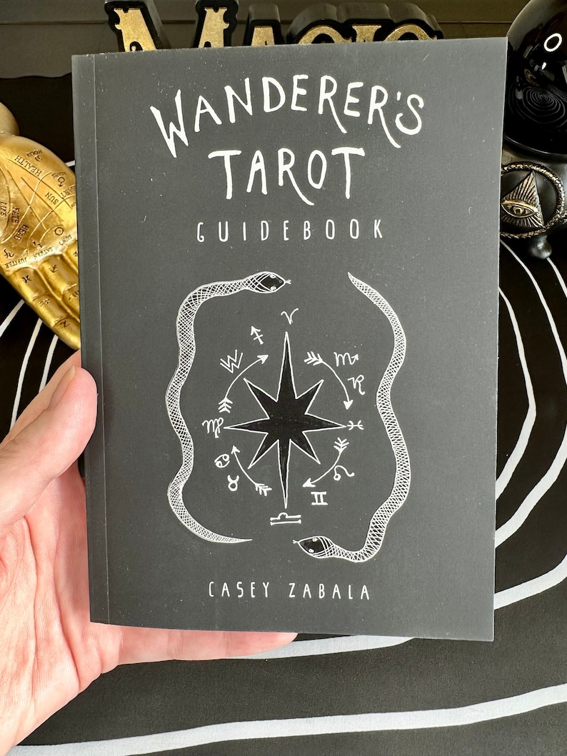 May include: A black guidebook with the title "Wanderer's Tarot Guidebook" in white text. The cover features a white illustration of a compass with a star in the centre and two snakes wrapped around it. The author's name, "Casey Zabala", is printed in white at the bottom.