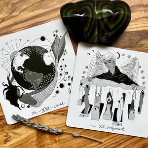 May include: Two tarot cards with black and white illustrations. One card depicts a mermaid with a globe, labeled "The - XXI - World." The other shows an angel above figures, labeled "~ XX judgement." A green, heart-shaped stone sits above the cards.