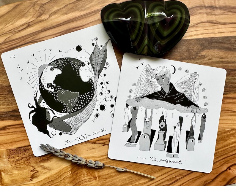 Może przedstawiać: Dwie karty tarota z czarno-białymi ilustracjami. Jedna karta przedstawia syrenę z globusem, zatytułowana "The - XXI - World". Druga przedstawia anioła nad postaciami, zatytułowana "~ XX judgement". Zielony kamień w kształcie serca leży nad kartami.