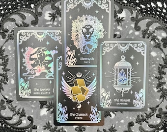 May include: Four tarot cards with holographic accents are displayed. The cards feature illustrations of a lion, a lantern, a crescent moon, and a winged cube. The cards are labeled "Strength," "The Hermit," "The Lovers," and "The Chariot."