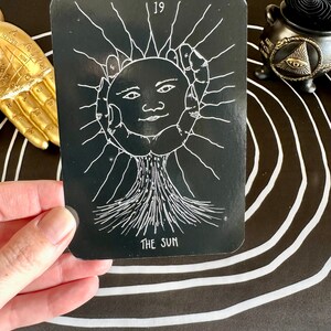 May include: A black tarot card with a white illustration of the sun, a face with a smile, and rays of light. The card number 19 and the words "The Sun" are printed in white on the card.