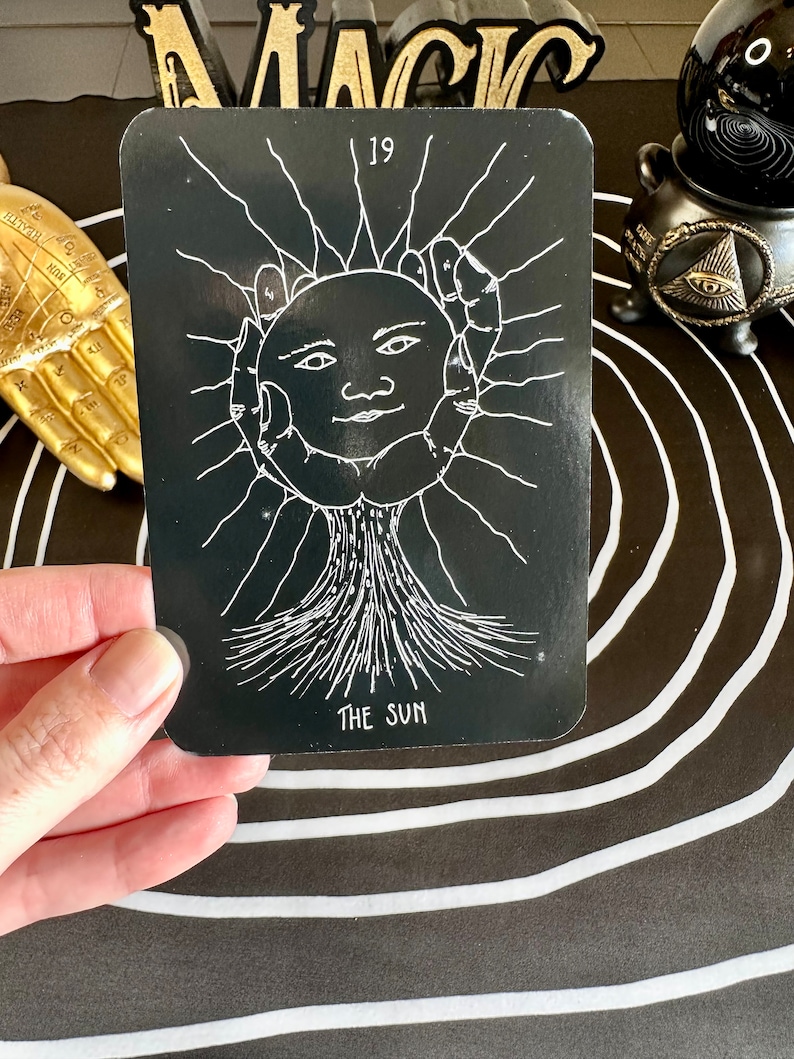 May include: A black tarot card with a white illustration of the sun, a face with a smile, and rays of light. The card number 19 and the words "The Sun" are printed in white on the card.
