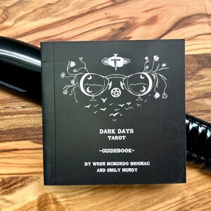 Może przedstawiać: Czarny podręcznik do tarota zatytułowany "Dark Days Tarot - Guidebook" z białymi ilustracjami i tekstem. Książka leży na drewnianej powierzchni z czarnym, cylindrycznym przedmiotem i czarnym przedmiotem w kształcie ostrza.