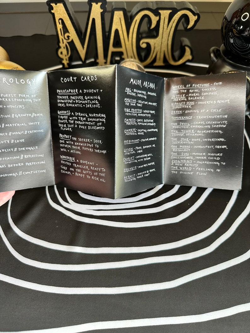May include: A set of black and silver tarot cards with gold lettering. The cards are folded to show the titles "Court Cards", "Major Arcana", and "Wheel of Fortune". The cards feature descriptions of each card's meaning.