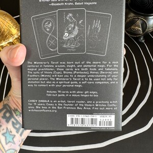 May include: A black box with a silver gilt edge containing a 78-card tarot deck. The box features a quote from Elizabeth Krohn, Sabat Magazine: "Inspired by nature and the cosmos. Wanderer's Tarot is a witchier deck with an air of the darkly feminine." The box also includes information about the creator, Casey Zabala, and the Modern Witches Confluence.