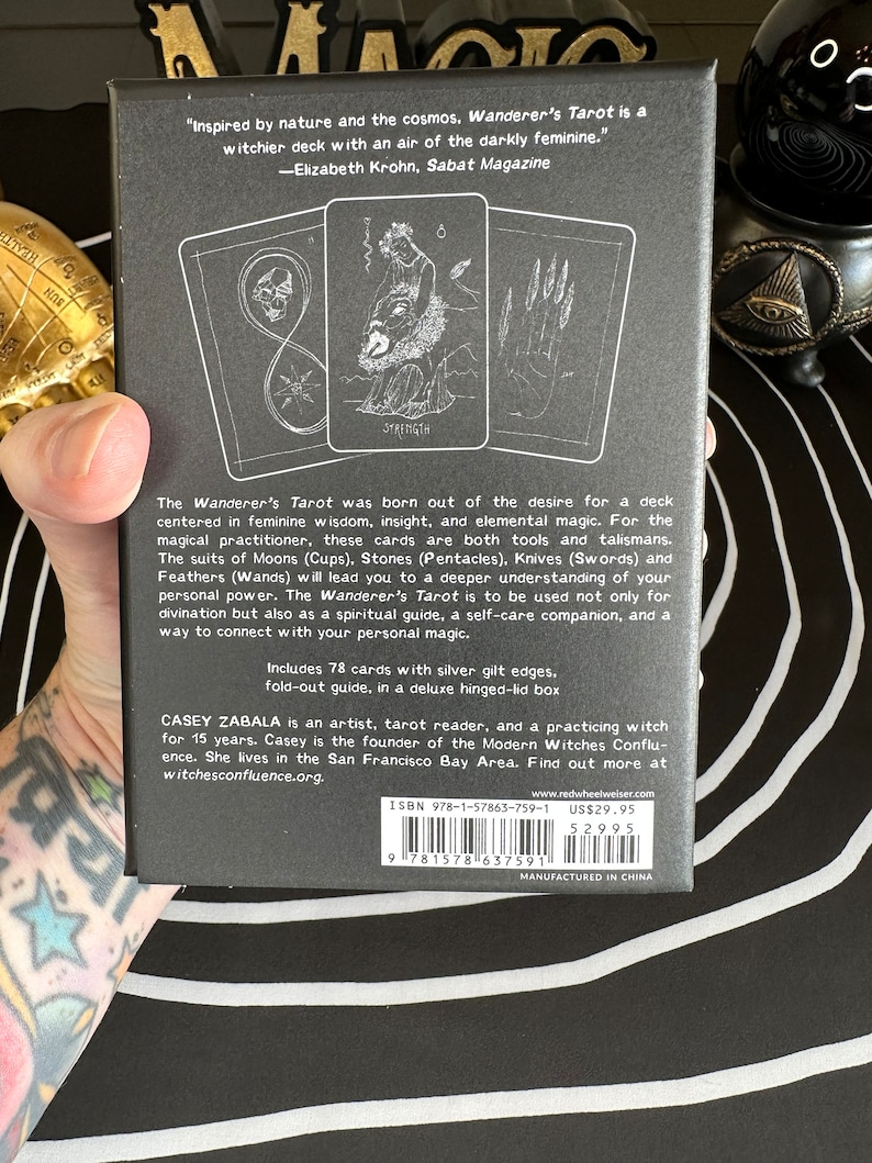 May include: A black box with a silver gilt edge containing a 78-card tarot deck. The box features a quote from Elizabeth Krohn, Sabat Magazine: "Inspired by nature and the cosmos. Wanderer's Tarot is a witchier deck with an air of the darkly feminine." The box also includes information about the creator, Casey Zabala, and the Modern Witches Confluence.