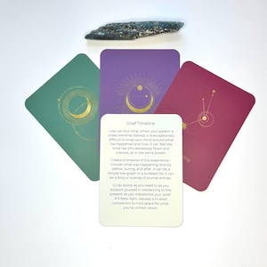 Puede incluir: Un conjunto de cartas de oráculo en tonos verdes, morados y burdeos, con diseños celestiales dorados. Una piedra oscura y alargada descansa sobre las cartas. Una carta muestra el texto: "Grief Timeline". Las cartas están dispuestas sobre una superficie blanca.