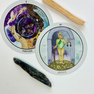 May include: Two circular tarot cards labelled Fortune and Justice, a wooden stick, and a dark blue crystal. The Fortune card features a purple galaxy centre and hands, while the Justice card depicts a figure with a moon head holding scales and a sword.