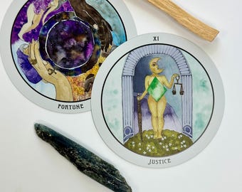 May include: Two circular tarot cards labeled Fortune and Justice, a wooden stick, and a dark blue crystal. The Fortune card features a purple galaxy center and hands, while the Justice card depicts a figure with a moon head holding scales and a sword.