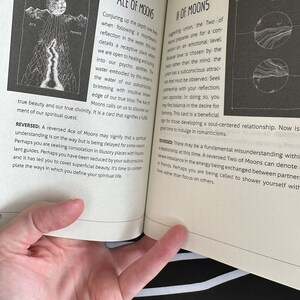 May include: A book page with a black and white illustration of a moon and a river. The text on the page reads "Ace of Moons" and "Two of Moons". The text describes the meaning of the cards in a tarot deck.