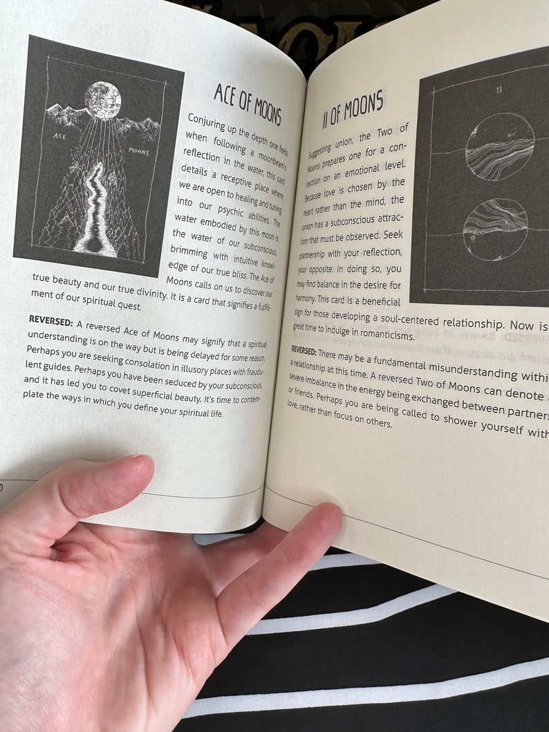 May include: A book page with a black and white illustration of a moon and a river. The text on the page reads "Ace of Moons" and "Two of Moons". The text describes the meaning of the cards in a tarot deck.