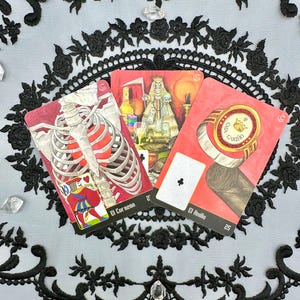 May include: Three tarot cards with red borders are displayed on a white and black lace background. The cards feature illustrations of a skeleton, a queen, and a ring. The card titles are in Spanish.