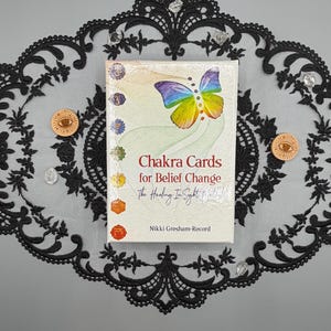 May include: A rectangular deck of Chakra Cards for Belief Change by Nikki Gresham-Record. The cover features a rainbow-colored butterfly, seven vertical chakra symbols, and the text The Healing In Sight Method. The deck is centered on a black lace mat.