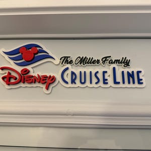 May include: A white sign with the words "The Miller Family" and "Disney Cruise Line" in black lettering. The Disney Cruise Line logo features a red Mickey Mouse head on a blue and white wave.